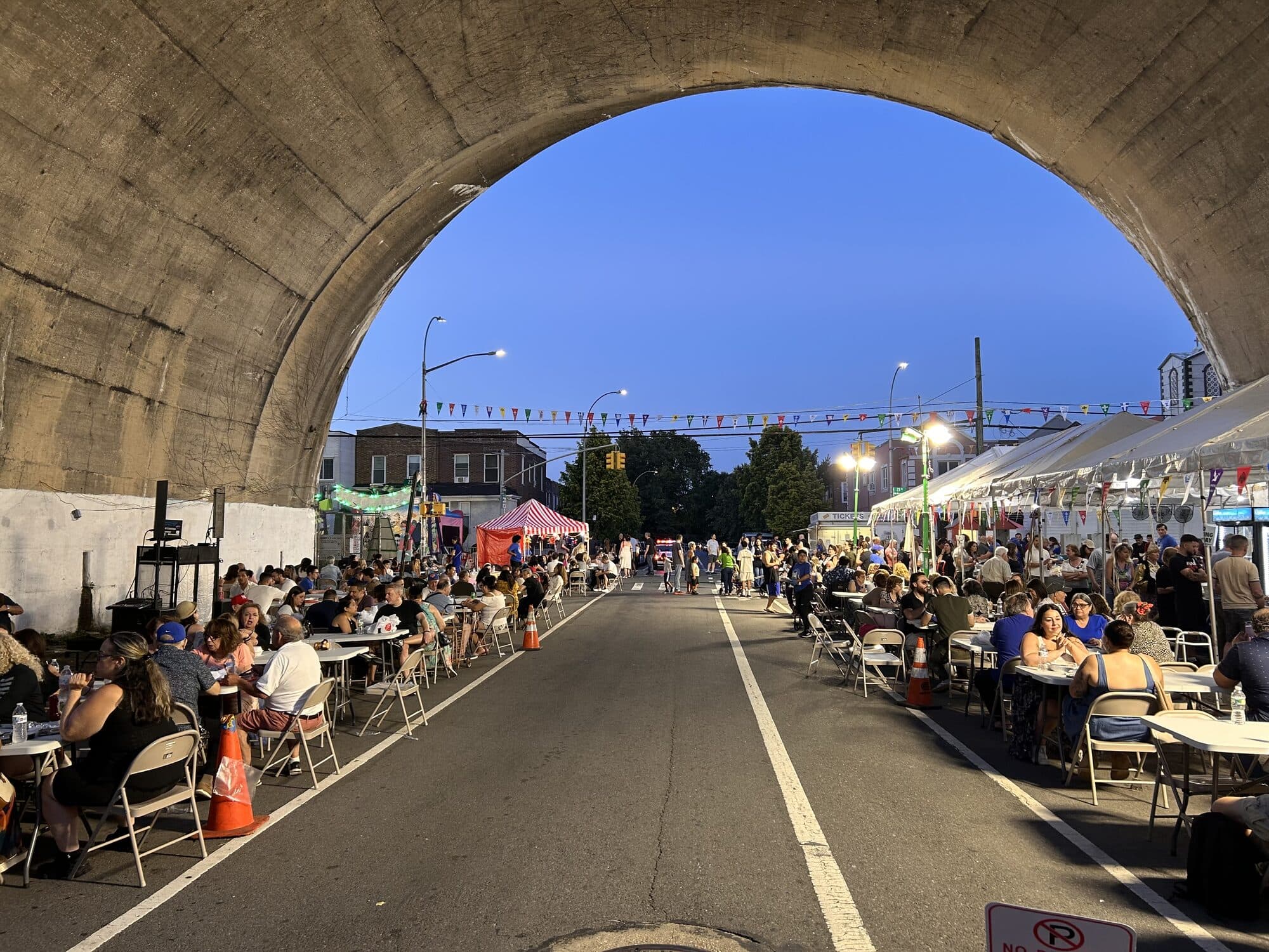 Festivalgoers fill long tables beneath the railroad viaduct on 23rd Avenue at dusk during the St. Irene Chrysovalantou panegyri